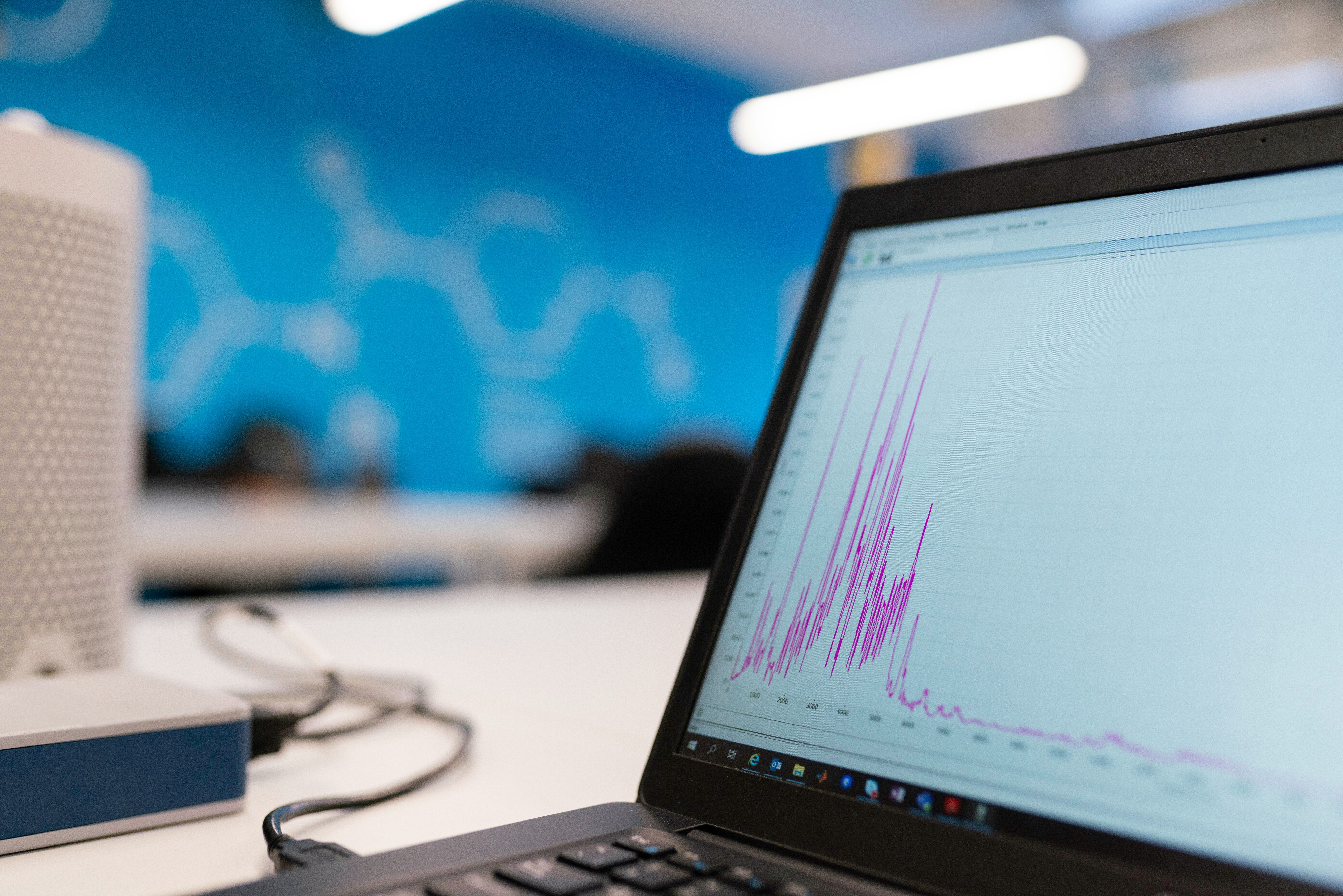 Softwarequalität: Ein Laptop auf einem weißen Schreibtisch zeigt auf seinem Bildschirm ein Diagramm mit magentafarbenen/violetten Ausschlägen, möglicherweise Datenanalyse oder Monitoring-Graphen. Im Hintergrund ist eine unscharfe blaue Wand mit grafischen Elementen zu sehen, und links im Bild befindet sich ein weißes Gerät mit einer perforierten Oberfläche, vermutlich ein Lautsprecher oder ein kleiner Server. Die Szene wirkt wie ein moderner Büro- oder Laborarbeitsplatz mit Datenvisualisierung.