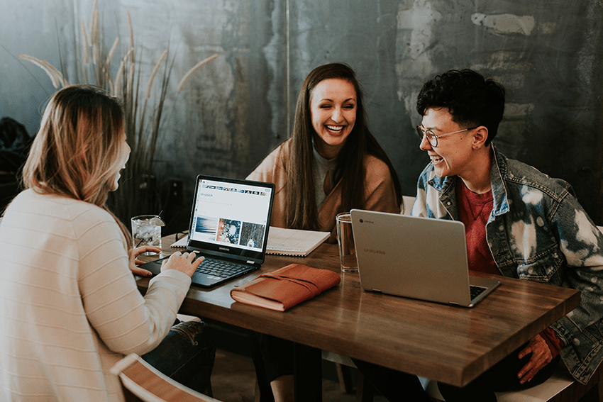 3 Personen sitzen mit Notebook und Notizbuch an einem Tisch und lachen