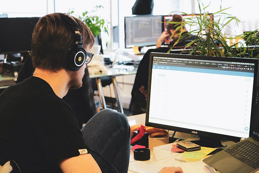 Mann sitzt mit Kopfhörern vor dem Monitor im Großraumbüro